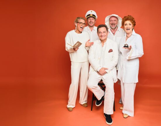 Darsteller von „Pesnion Schöller“ in ihren weißen Kostümen vor orangem Hintergrund